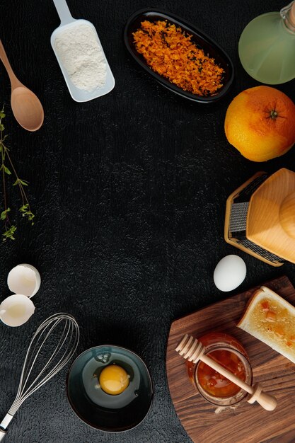 Top view of open flour egg orange lime juice jam bread slice orange zest with whisk spoon and grater with egg shell on black background