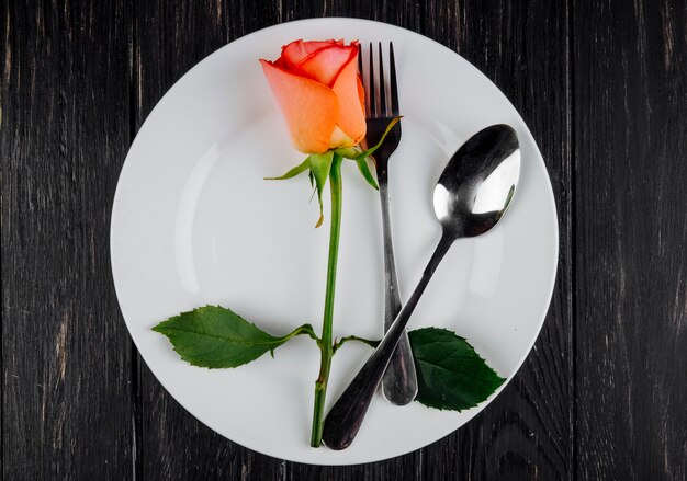 Top view of orange color rose with spoon and fork on a white plate on dark wooden background