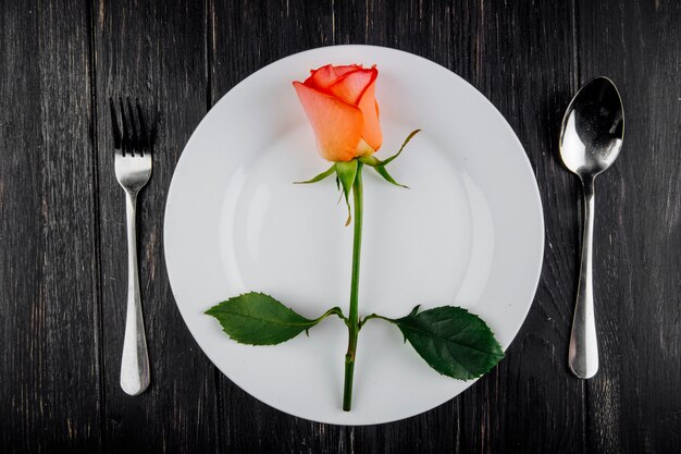 暗い背景の木の白い皿にスプーンとフォークでオレンジ色のバラのトップビュー