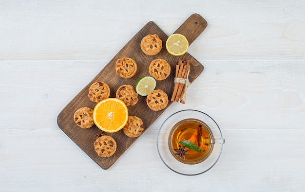 Free photo top view an orange, lime, cookies and cinnamon in cutting board with a cup of tea on white surface