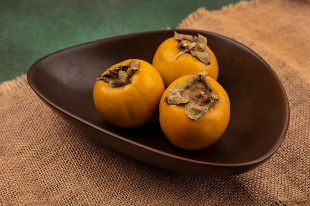 Top view of orange persimmon fruits on a bowl on a sack cloth on a green wall