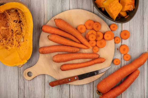Top view of orange root vegetable carrots on a wooden kitchen board with chopped carrots with knife with half pumpkin on a grey wooden surface