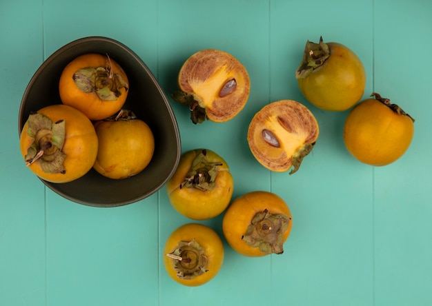 Foto gratuita vista dall'alto dei frutti di cachi tondeggiante arancione su una ciotola con frutti di cachi isolati su un tavolo di legno blu