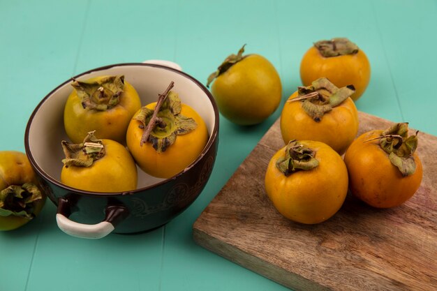 top view of orange roundish persimmon fruits on a wooden kitchen board with persimmon fruits on a bowl on a blue wooden table