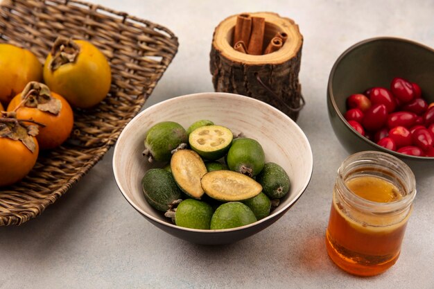 Top view of organic feijoas on a bowl with persimmons on a wicker tray with cinnamon sticks with cornelian cherries on a bowl with honey on a glass jar on a grey background