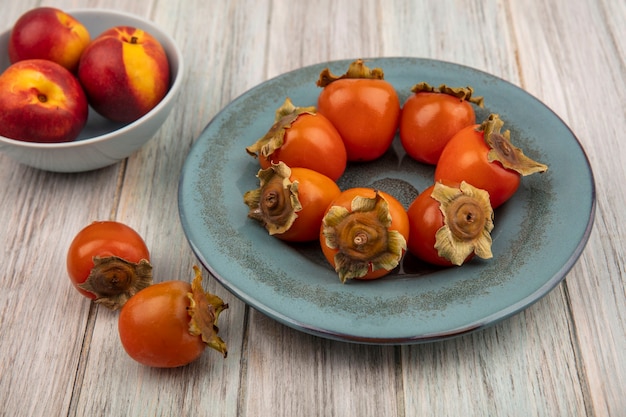 Top view of organic peaches on a bowl with soft fresh persimmons on a plate on a grey wooden background