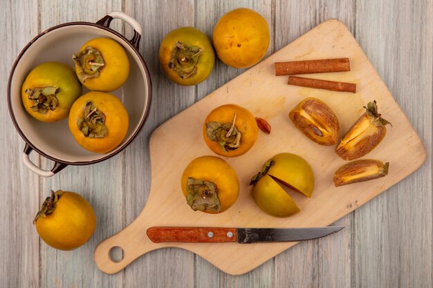 top view of organic persimmon fruits on a wooden kitchen board with cinnamon sticks with knife on a grey wooden table