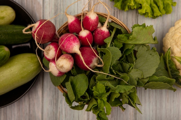 Top view of organic radishes on a bucket with cucumbers and zucchinis on a plate with lettuce and cauliflower isolated on a grey wooden surface