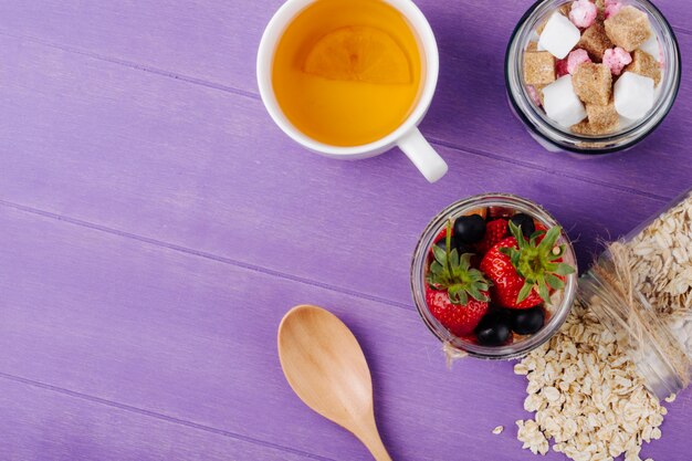 Top view of overnight oats with fresh strawberries blueberries yogurt and nuts in a glass jar with a cup of green tea on purple wooden surface with copy space