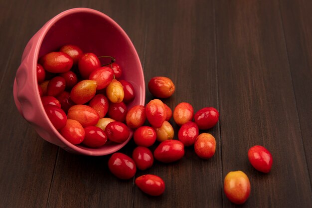 Top view of pale red sour cornelian cherries falling out of a bowl on a wooden surface