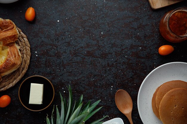 검은 배경에 파인애플 잎이 있는 팬케이크 잼 버터 금귤 크루아상 밀가루의 상위 뷰