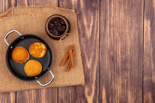 Top view of pancakes in pan and jar of strawberry jam with cinnamon on sackcloth on wooden background with copy space