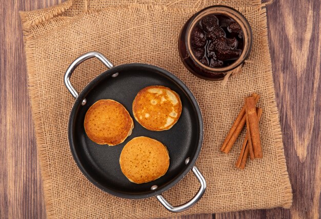 Foto gratuita vista dall'alto di frittelle in padella e vasetto di marmellata di fragole con cannella su tela di sacco su fondo in legno