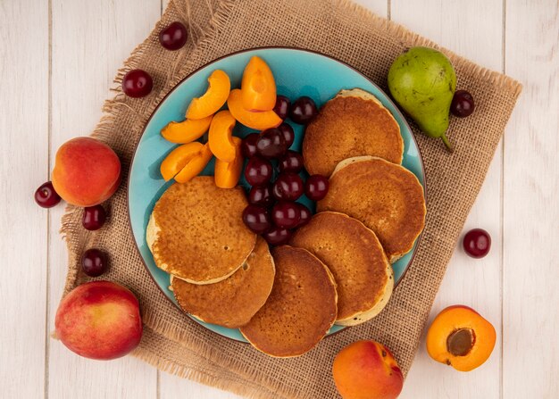 Top view of pancakes with cherries and apricot pieces in plate and apricots cherries pear on sackcloth and on wooden background