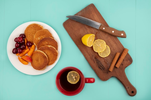 Top view of pancakes with cherries and apricot slices in plate and cup of coffee with lemon slices and cinnamon with knife on cutting board on blue background