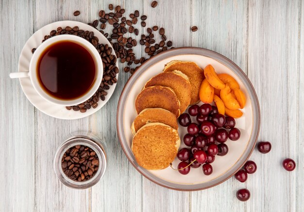 Top view of pancakes with cherries and apricot slices in plate and cup of tea with coffee beans on saucer and in jar on wooden background