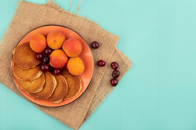 Top view of pancakes with cherries and apricots in plate on sackcloth on blue background with copy space