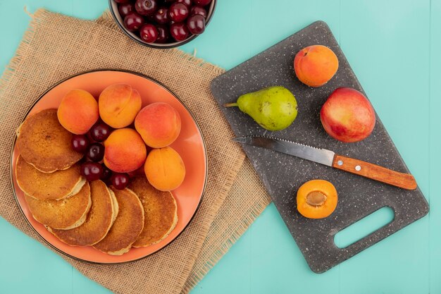 Top view of pancakes with cherries and apricots in plate on sackcloth with bowl of cherries and pear peach apricot with knife on cutting board on blue background