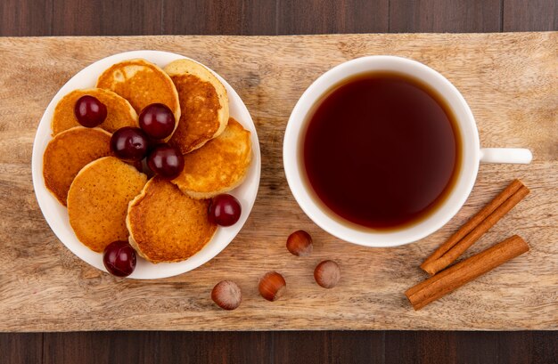 Top view of pancakes with cherries in plate and cup of tea with cinnamon and nuts on cutting board on wooden background