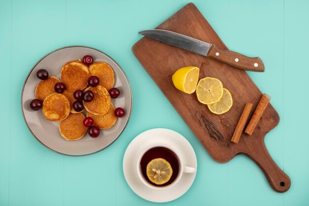 Top view of pancakes with cherries in plate and cup of tea with lemon slices and cinnamon with knife on cutting board on blue background