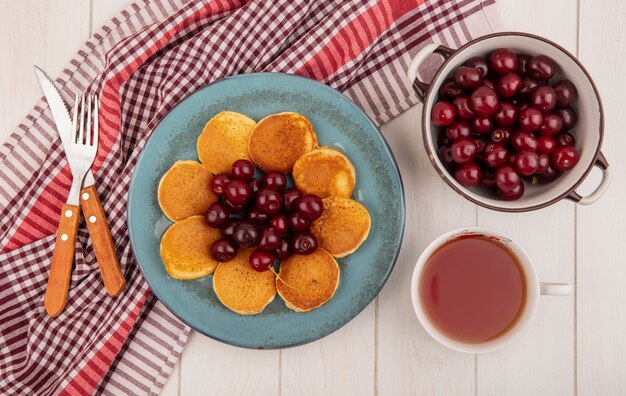 Top view of pancakes with cherries in plate and fork knife on plaid cloth and bowl of cherries with cup of tea on wooden background