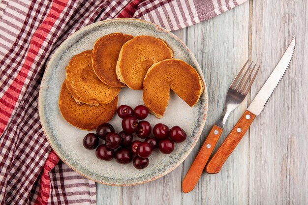 Top view of pancakes with cherries in plate on plaid cloth and fork knife on wooden background