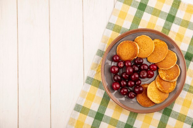 Top view of pancakes with cherries in plate on plaid cloth and wooden background with copy space