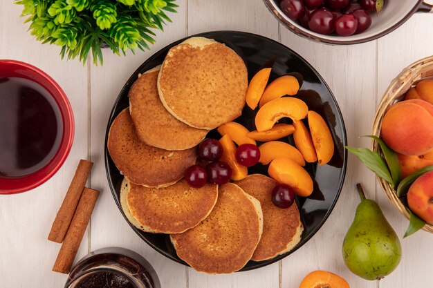 Vista dall'alto di frittelle con ciliegie e albicocche a fette nel piatto e tazza di caffè con marmellata di cannella pera e albicocche su fondo di legno