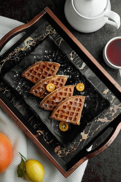 Top view of pancakes with kumquat slices in plate and orange lemon on cloth with teapot and cup of tea on black background