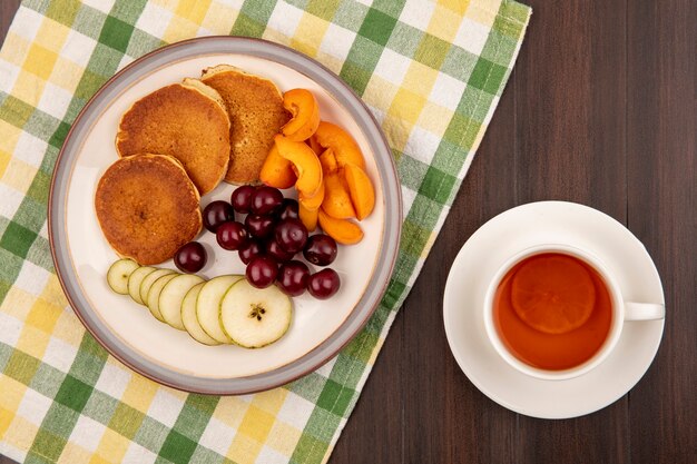 木製の背景にお茶のカップと格子縞の布の上の梨とアプリコットスライスとチェリーのプレートのパンケーキのトップビュー