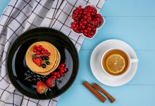 Top view of pancakes with red and black currants and strawberries on a black plate with a cup of tea on a blue surface