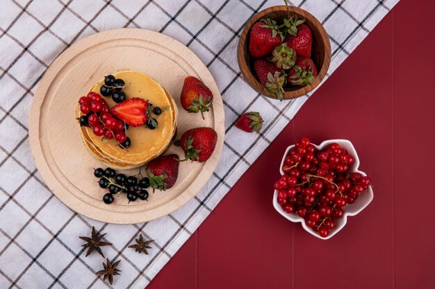 Top view pancakes with red and black currants and strawberries on a towel on a red background