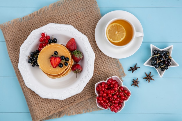 Top view of pancakes with red and black currants and strawberries with a cup of tea on a blue surface