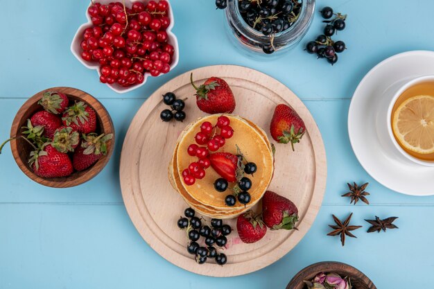 Top view of pancakes with red and black currants and strawberries with a cup of tea on a blue surface