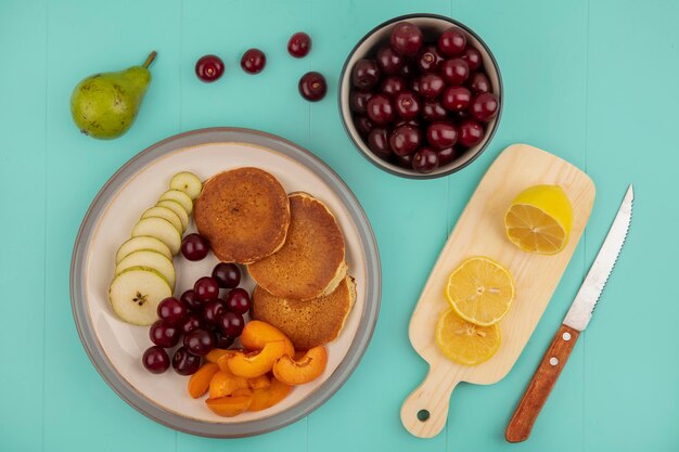 Top view of pancakes with sliced apricot and pear and cherries in plate with sliced lemon on cutting board and bowl of cherries with knife on blue background