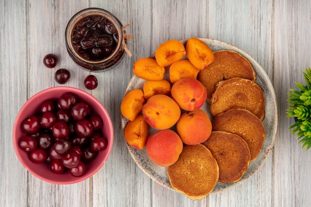 Top view of pancakes with whole and sliced apricots in plate and bowl of cherries with strawberry jam on wooden background