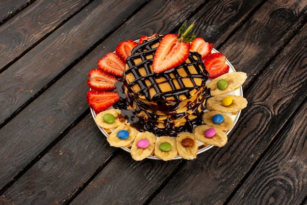 Foto gratuita vista dall'alto pancake deliziosi con fragole rosse a fette e banane all'interno del piatto bianco sul pavimento rustico in legno marrone