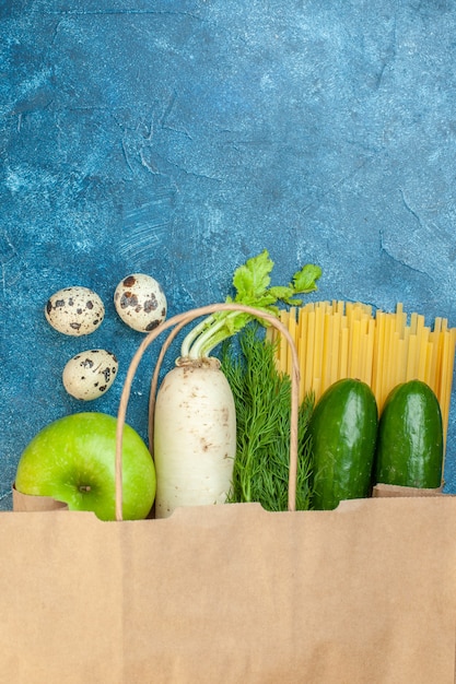 Top view paper shopping bag eggs radish cucumber spaghetti dill apple on blue table