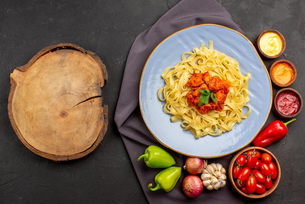 Вид сверху макароны на скатерти синяя тарелка аппетитной пасты миска из помидоров, соусы, чеснок, лук, шарик, перец на фиолетовой скатерти и деревянная доска на столе