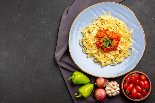 Top view pasta on tablecloth plate of appetizing pasta next to the bowl of tomatoes garlic onion green ball pepper on the purple tablecloth on the table