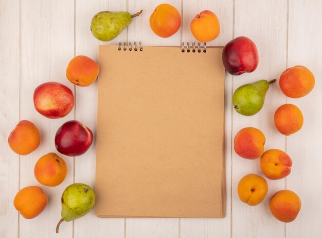 Вид сверху на узор из фруктов в виде абрикоса, персика и груши вокруг блокнота на деревянном фоне с копией пространства