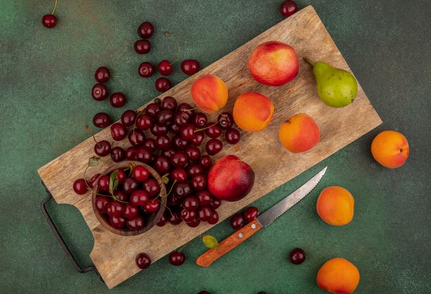 Top view of pattern of fruits as apricots peaches pear cherries with bowl of cherry on cutting board with knife on green background