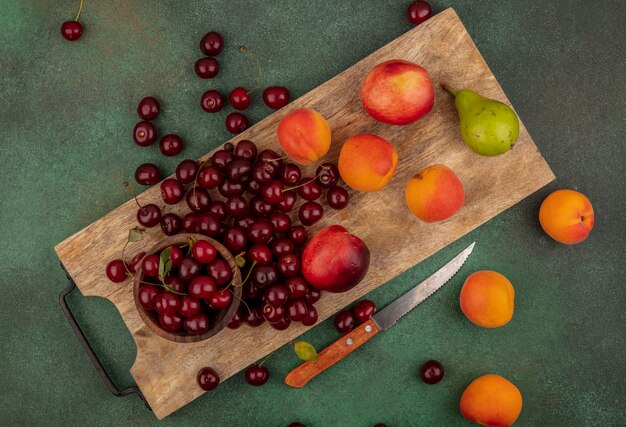 緑の背景にナイフでまな板の上のチェリーのボウルとアプリコット桃梨サクランボとして果物のパターンのトップビュー