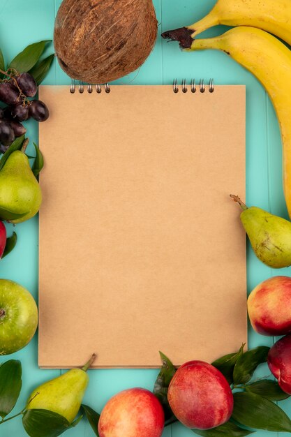 Top view of pattern of fruits as coconut pear peach grape banana apple around note pad on blue background with copy space