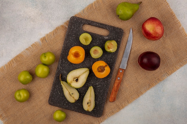Top view of pattern of fruits as half cut pear apricot plum on cutting board and peach pear plum with knife on sackcloth on white background