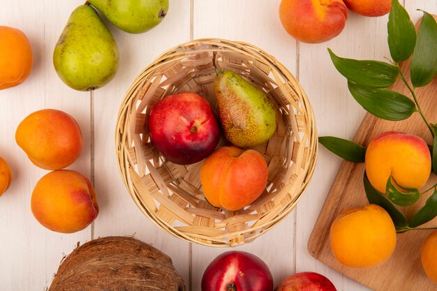 Top view of pattern of fruits as peach pear apricot in basket and peaches on cutting board with pears and coconut on wooden background