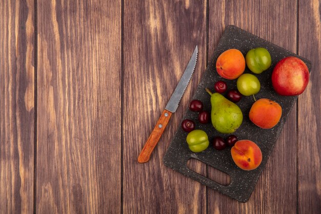 Top view of pattern of fruits as peach plums apricots pear cherries on cutting board with knife on wooden background with copy space