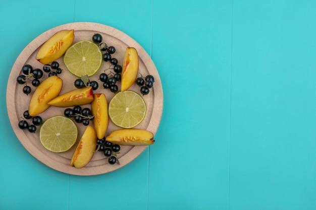 Top view of peach slices with blackcurrant and lime slices on a wooden tray on a blue surface