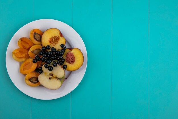 Top view of peach with apricots and an apple with black currants on a plate on a light blue surface