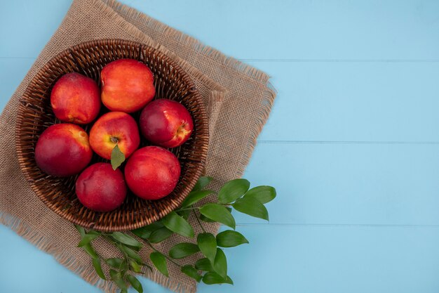 Top view of peaches in a basket with leaf branches on a beige napkin on a light blue surface